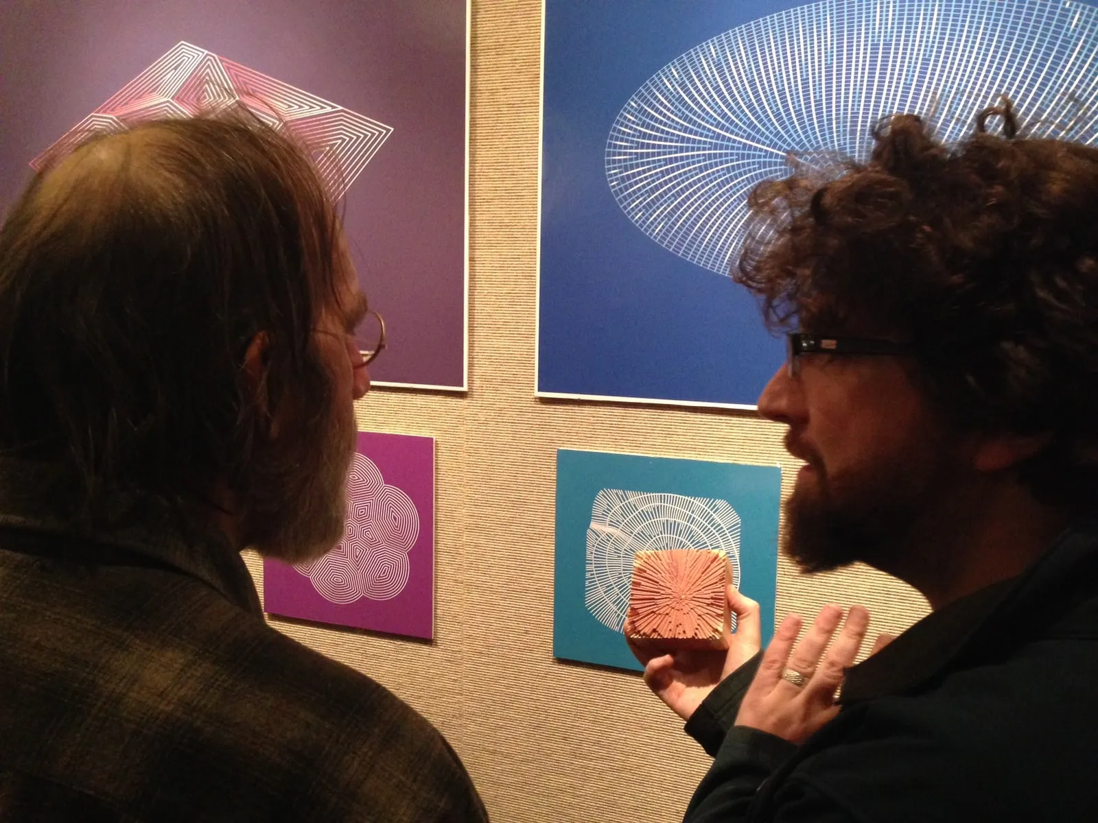 Two visitors at the opening reception examining framed line-pattern prints on coloured panels, one holding a small routed wooden disc with radiating lines