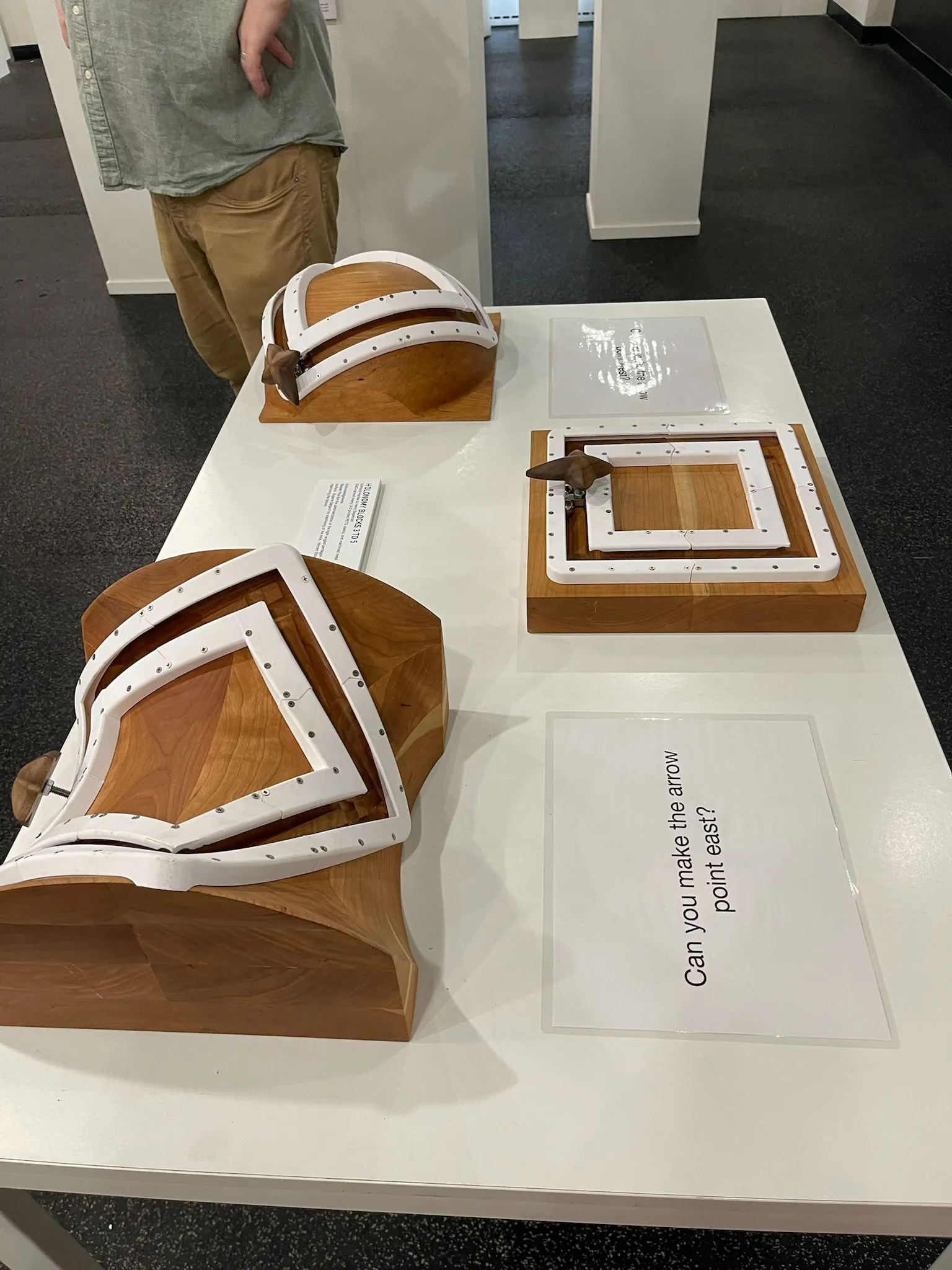 Three boards on a white table at MoMath (sphere, flat, and saddle), each with white metal tracks and a wooden arrow, with a prompt card reading 'Can you make the arrow point east?'