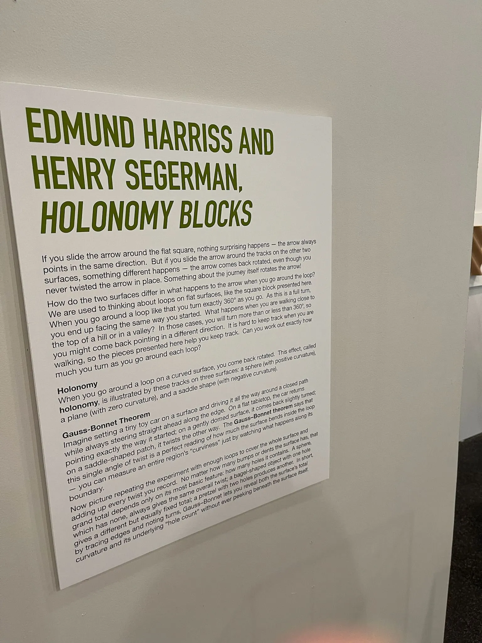 Exhibition wall text: "Edmund Harriss and Henry Segerman, Holonomy Blocks" with explanatory text about holonomy and the Gauss-Bonnet theorem