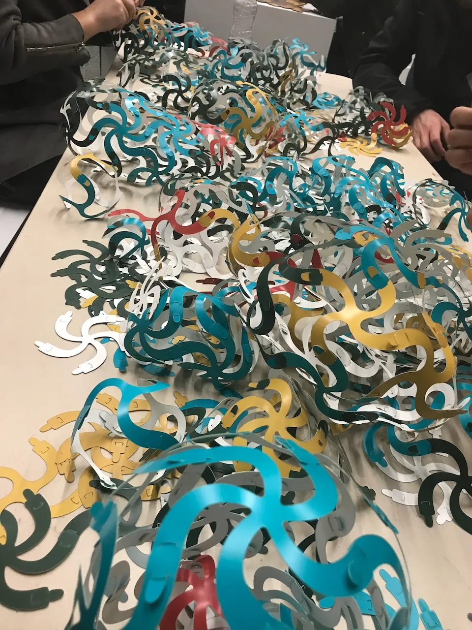 Hundreds of Curvahedra pieces spread across a long table ready for assembly,  teal, gold, red, white, and dark pieces