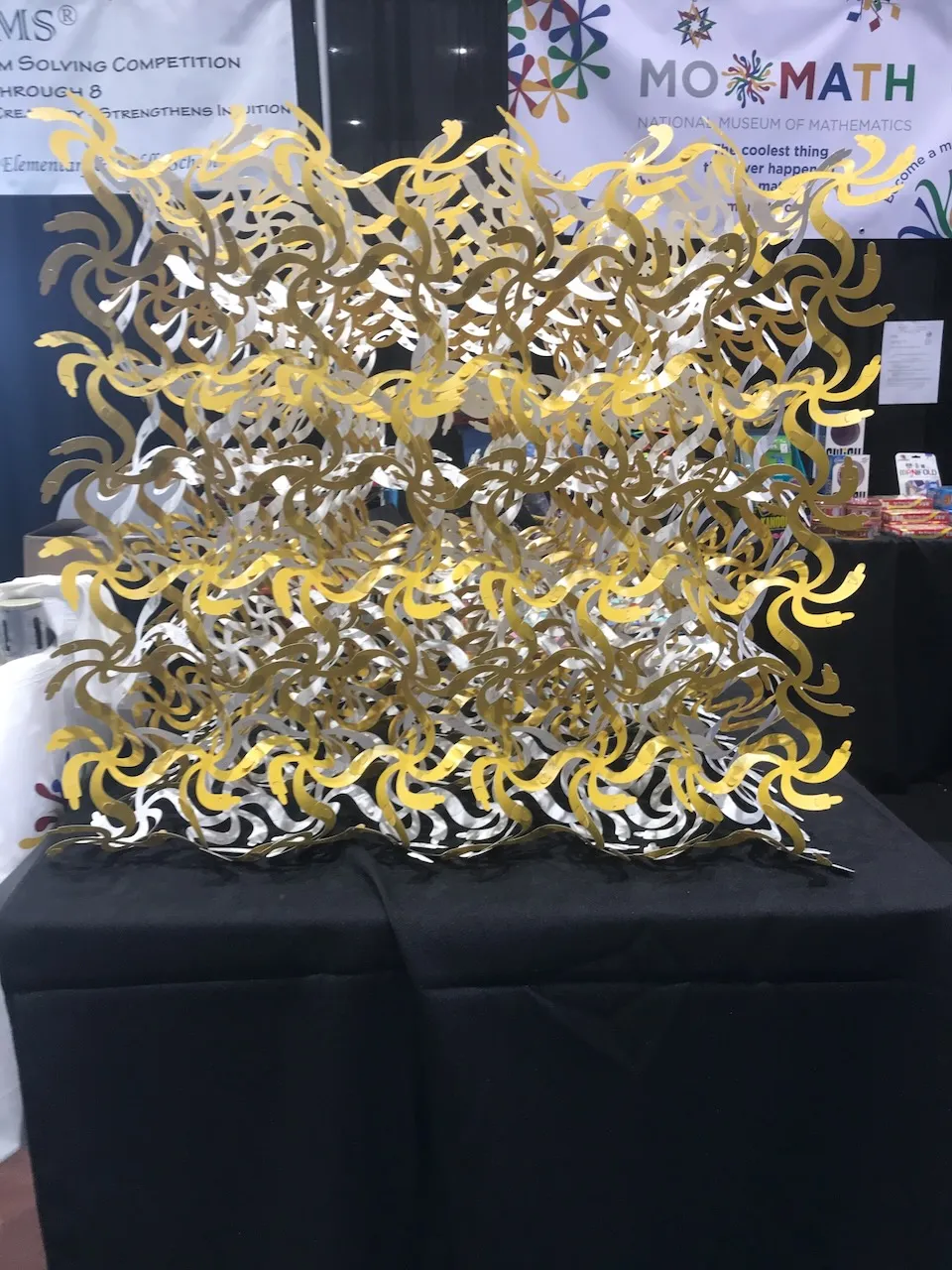 A large flat Curvahedra surface in gold and silver on a black table at the MoMath booth at a mathematical sciences conference