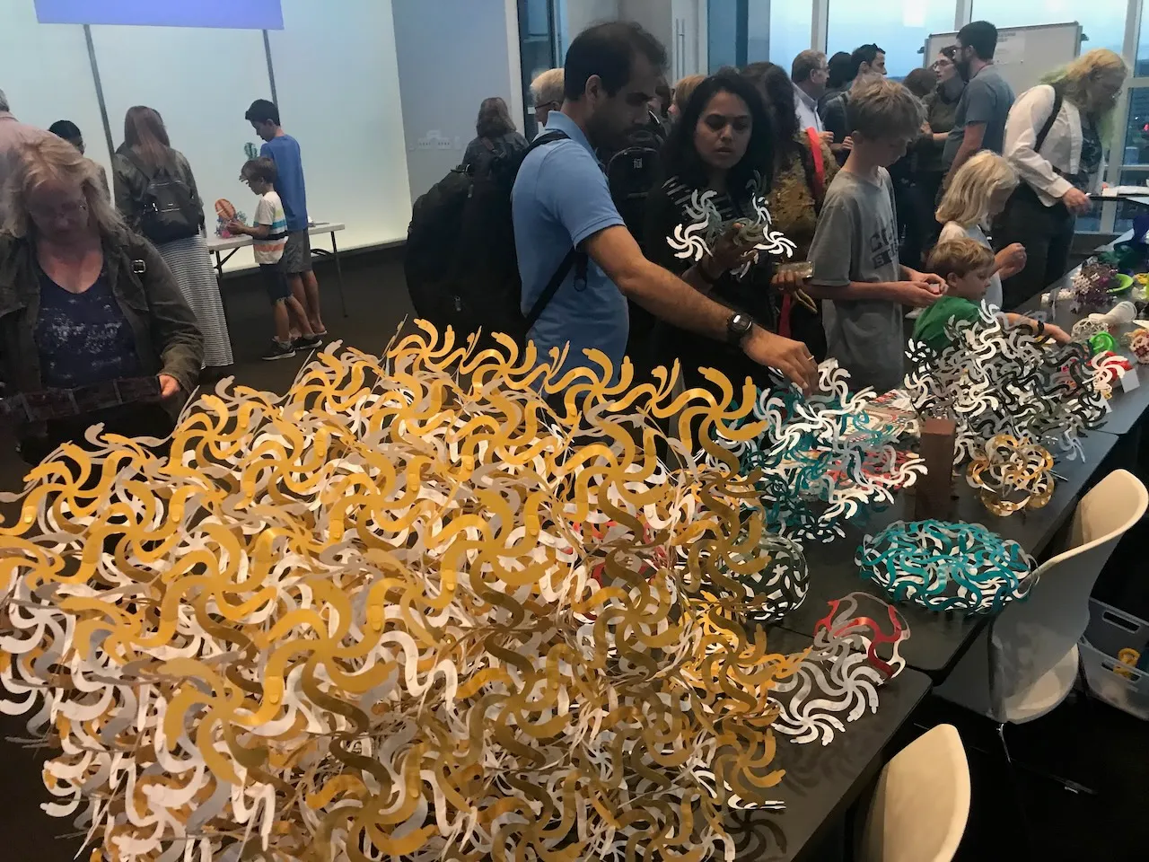 Visitors exploring Curvahedra pieces at a public event, a table covered in gold and white pieces with assembled forms at the far end