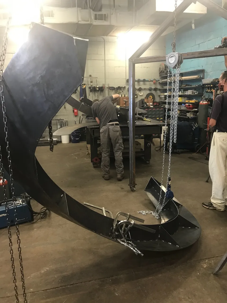 Two large curved steel elements suspended by chain hoists in the fabrication shop, a welder working at the bench behind them