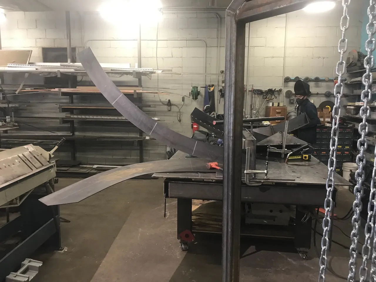Curved steel elements clamped to a welding table mid-assembly, welder in the background, chain hoist hanging to the right