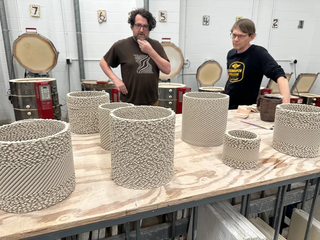 Edmund Harriss and Vincent Edwards in the ceramics studio examining the unfired clay-printed vessels, kilns visible behind them