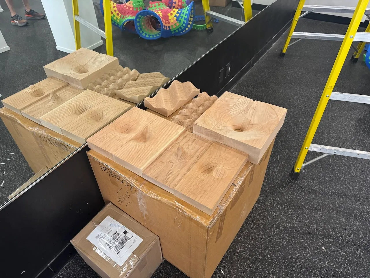 Geodesic Blocks and boards fresh from shipping, several carved wooden pieces stacked on cardboard boxes on the gallery floor