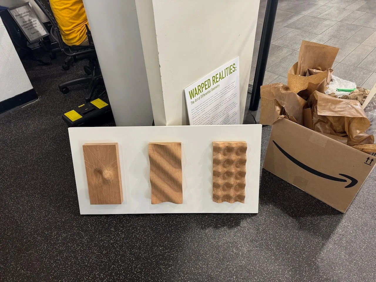 A Geodesic Boards panel on the floor next to unpacking materials, with the Warped Realities exhibition sign propped behind it