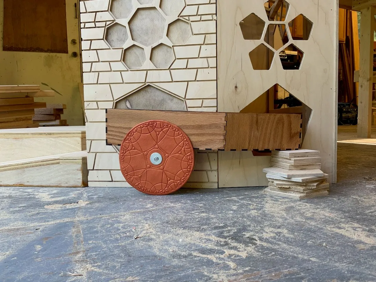 The terracotta Apollonian cart wheel leaning against the Bakery base in the workshop; Voronoi floor tile stack to the right