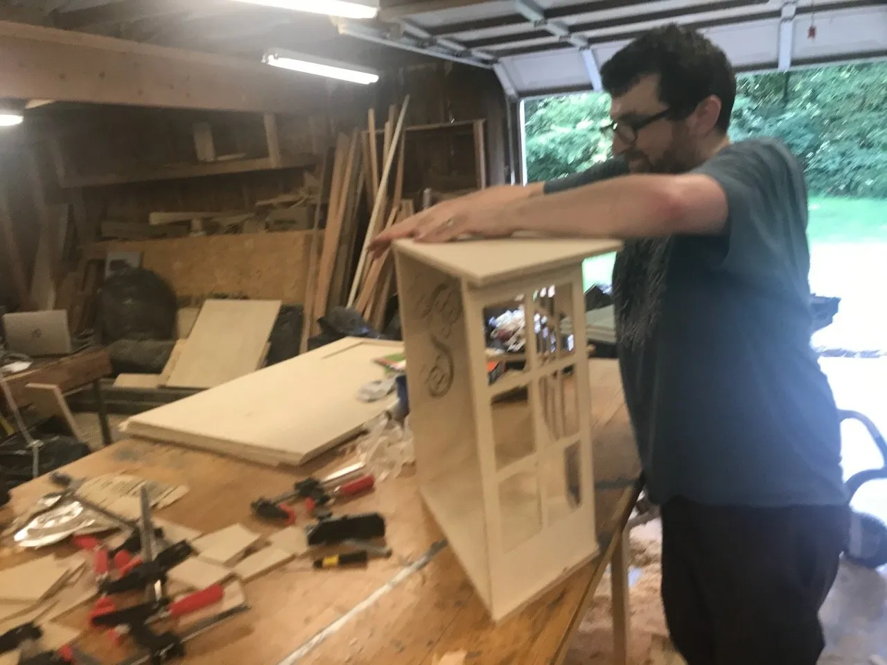 Edmund Harriss in the garage lifting the nearly finished Conway's Curios shop; the wave-pattern railing and painted exterior visible, clamps and workshop tools on the bench behind him