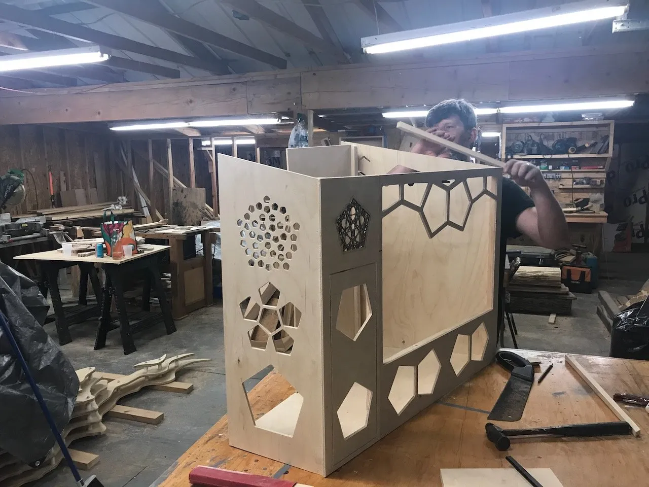 Edmund assembling the large heptagonal base structure in the workshop; plywood walls with circle-packing and pentagon cutout patterns, collaborator peering over the top edge, diagonal bracing visible