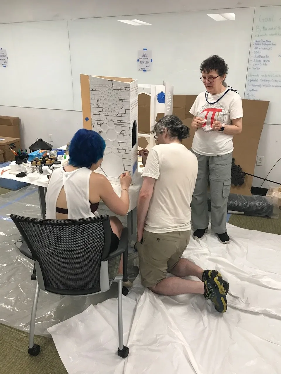 Three collaborators painting the Bakery brick wall white at the Duke assembly week, a woman in a blue top, a man kneeling, and a woman in a pi t-shirt standing, the oven arch facade visible