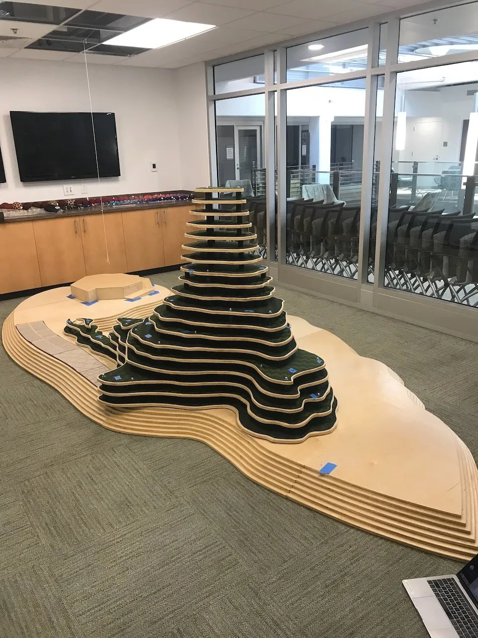 The Integral Hill fully assembled in a Duke University conference room; stacked CNC-cut contour layers in dark green fabric and pale wood, rising from the wavy plywood landscape base, the room's TV screen and oak cabinets visible