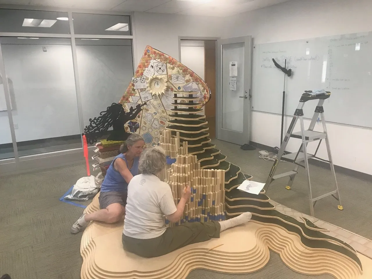 Two collaborators seated on the floor fitting wooden bead columns into the Integral Hill at the Duke assembly; the large topographic mountain rising behind them, the OctoPi illustration mural and a standing flute-girl silhouette visible