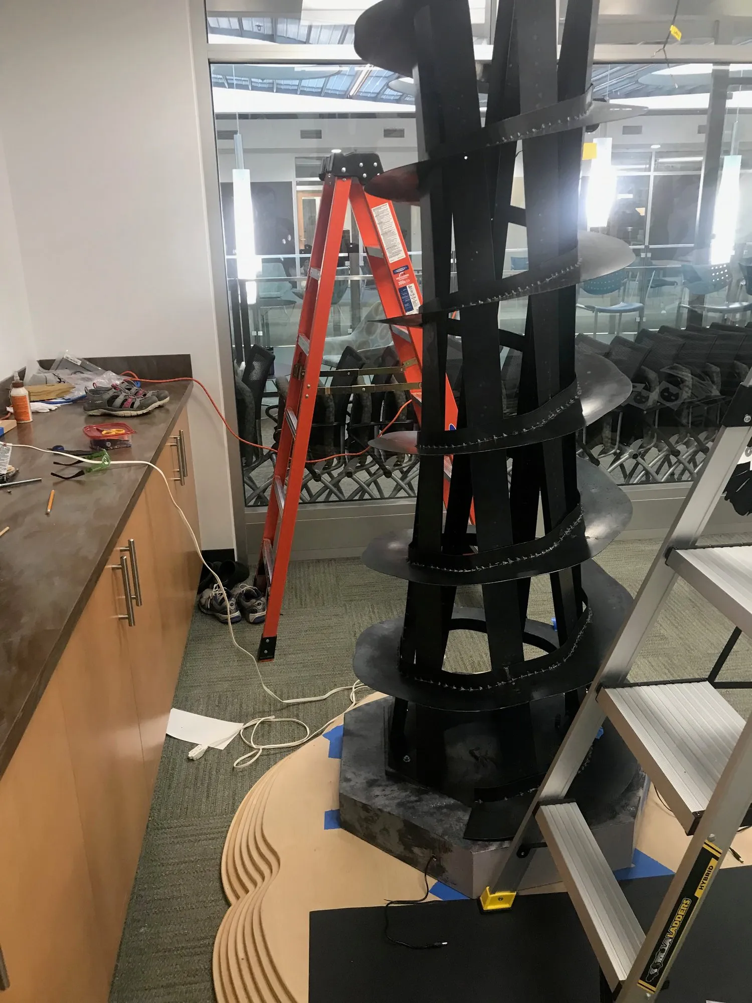 Lighthouse tower being assembled at Duke University with an orange stepladder leaning against it; a collaborator working near the top, the helical structure reaching the conference room ceiling tiles