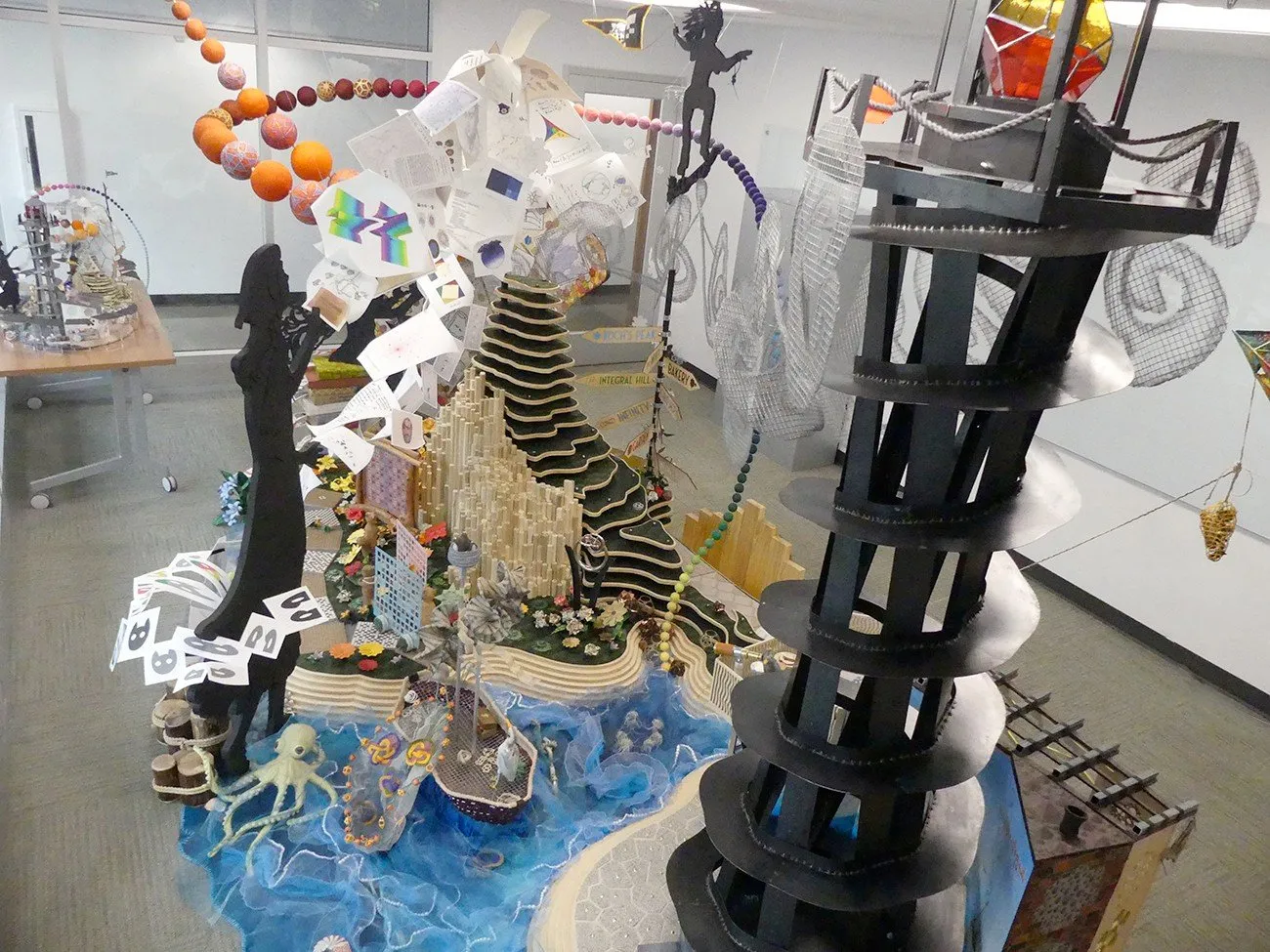 Overhead view of the complete Mathemalchemy installation laid out in a Duke conference room during the assembly week; Integral Hill, Lighthouse, Cavalcade arc, and all silhouette figures visible from above at an intermediate assembly stage