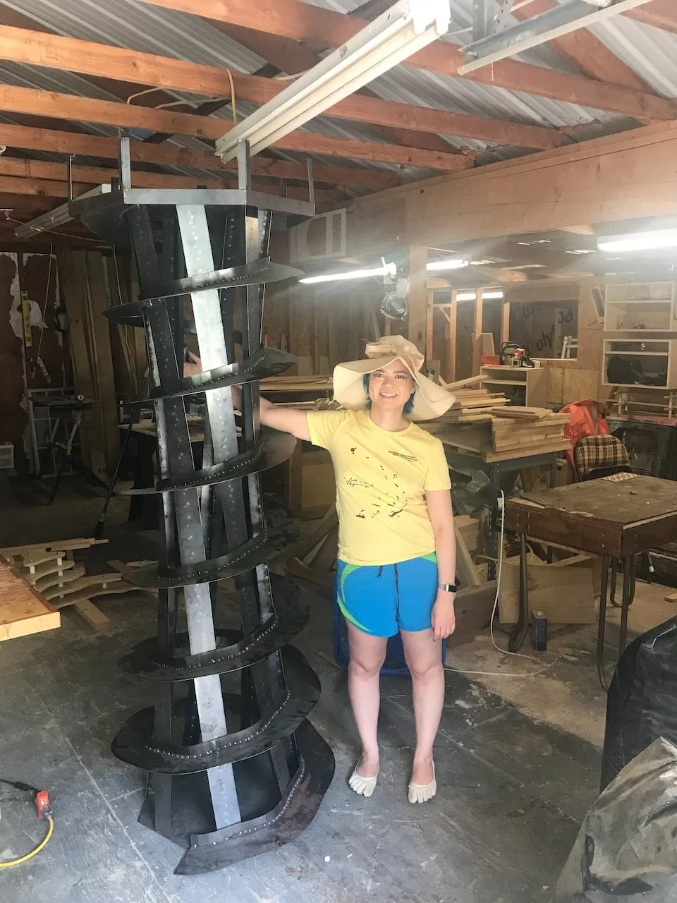 A woman in a yellow t-shirt and wide-brimmed hat posing beside the completed Lighthouse tower in her home garage workshop; the helical steel structure nearly reaches the ceiling, spiral staircase and landing plates clearly visible