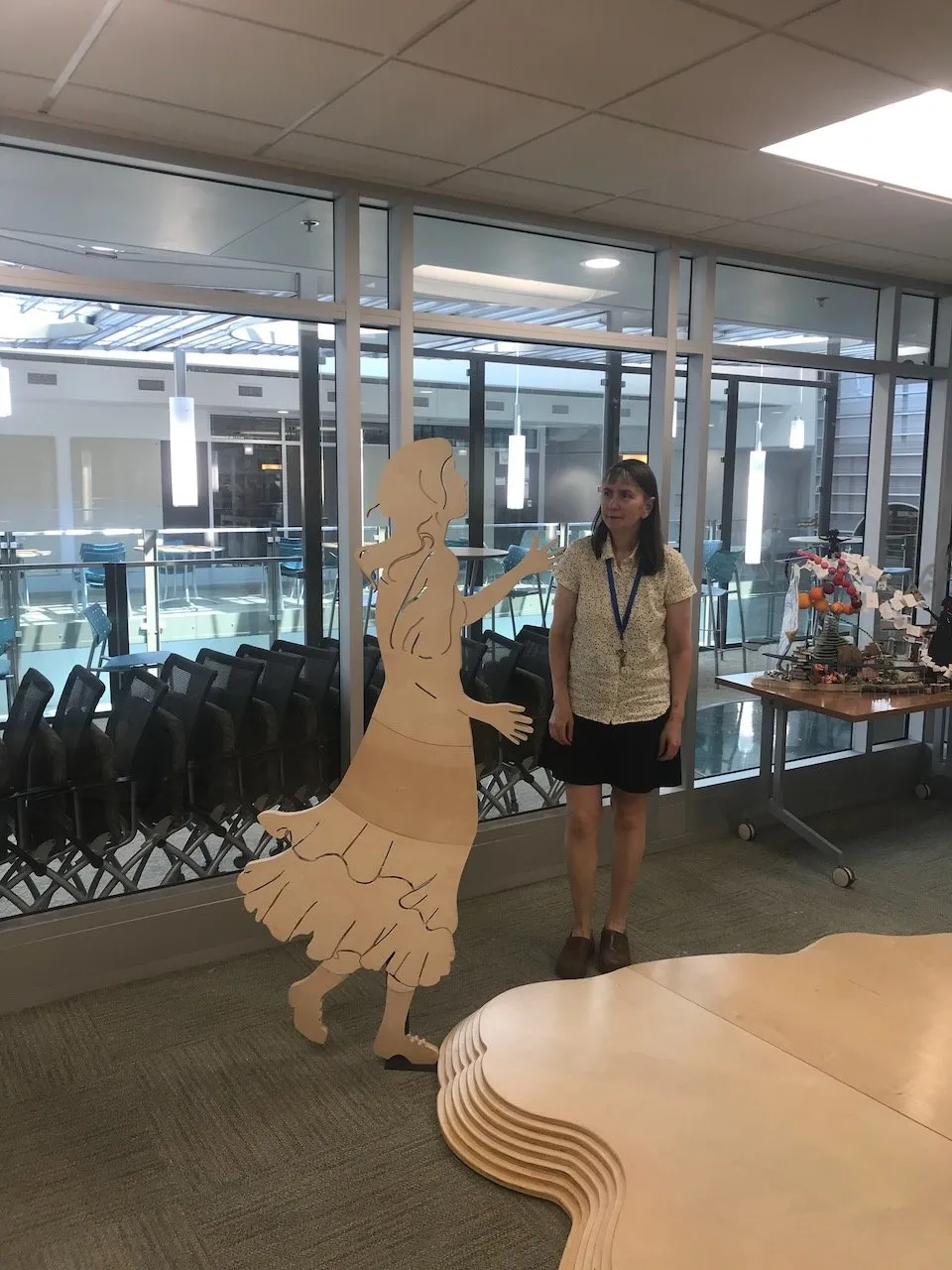 A woman collaborator in a white top and lanyard standing beside the assembled flute-girl figure in a Duke University conference room; the Integral Hill contour sections visible on the floor behind