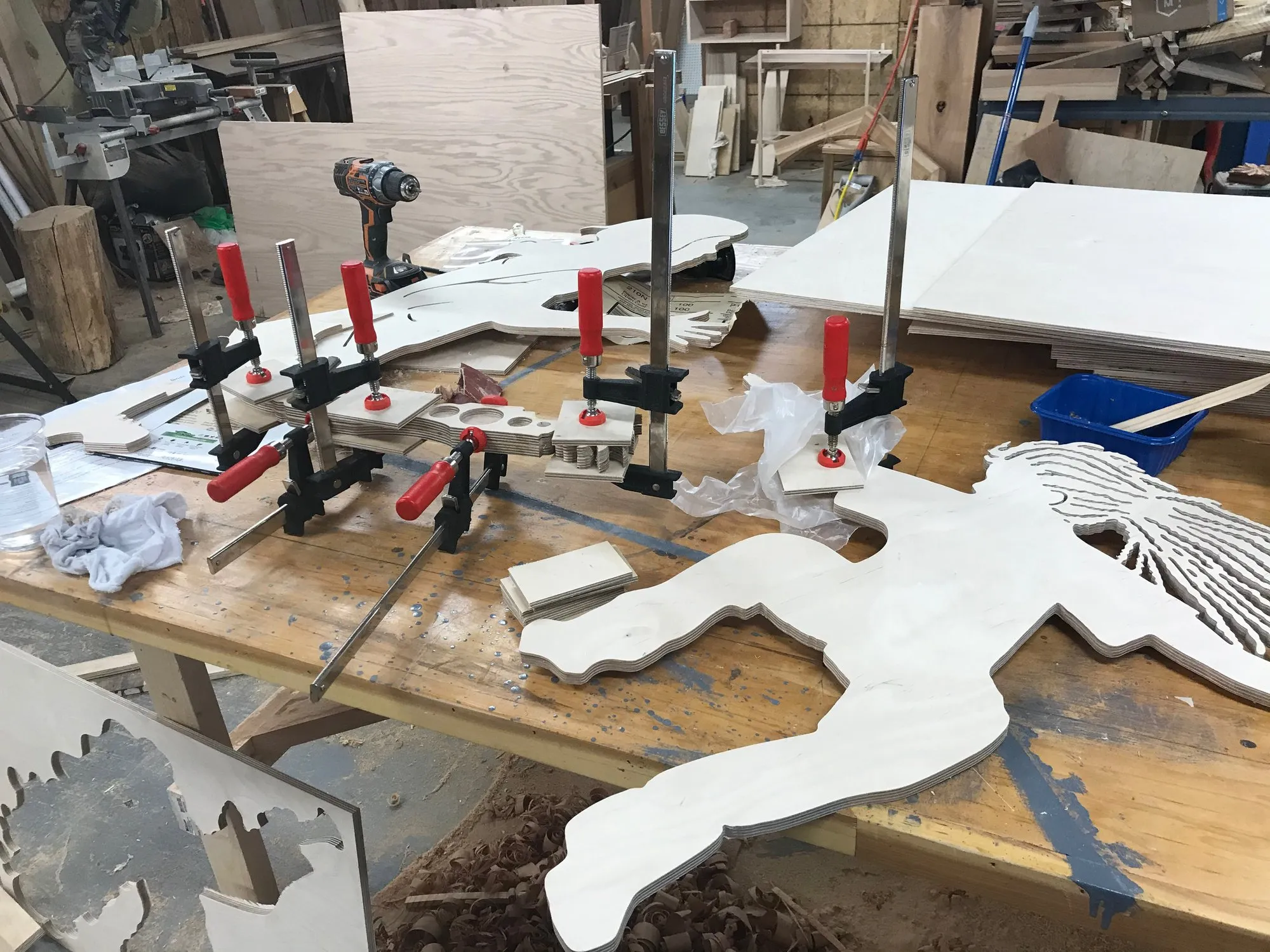 Workshop bench at the Duke assembly week; multiple white-primed plywood silhouette sections, clamps everywhere, a busy collaborative working session