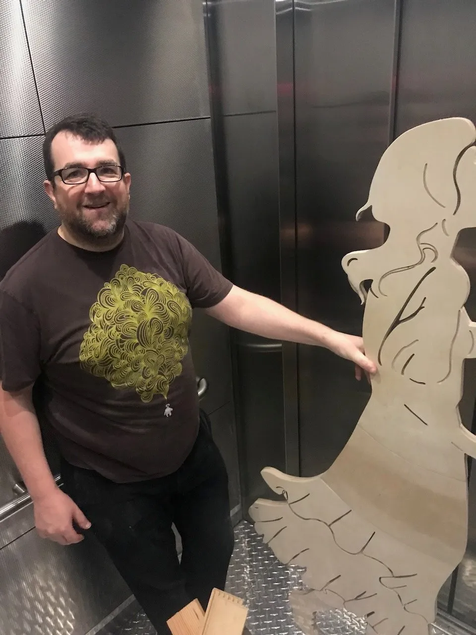 Edmund Harriss in a metal elevator or goods lift with the fully assembled flute-girl figure; his brown fractal-pattern t-shirt and glasses visible, the flowing plywood hair of the figure reaching above the lift door