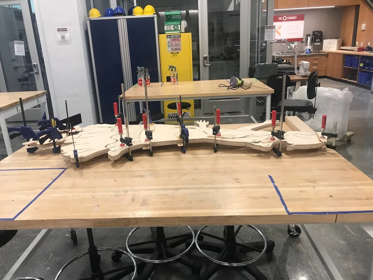 University fabrication lab; a full-length plywood silhouette figure being glued and laminated with many clamps spread across butcher-block tables, the multi-section body clamped into alignment