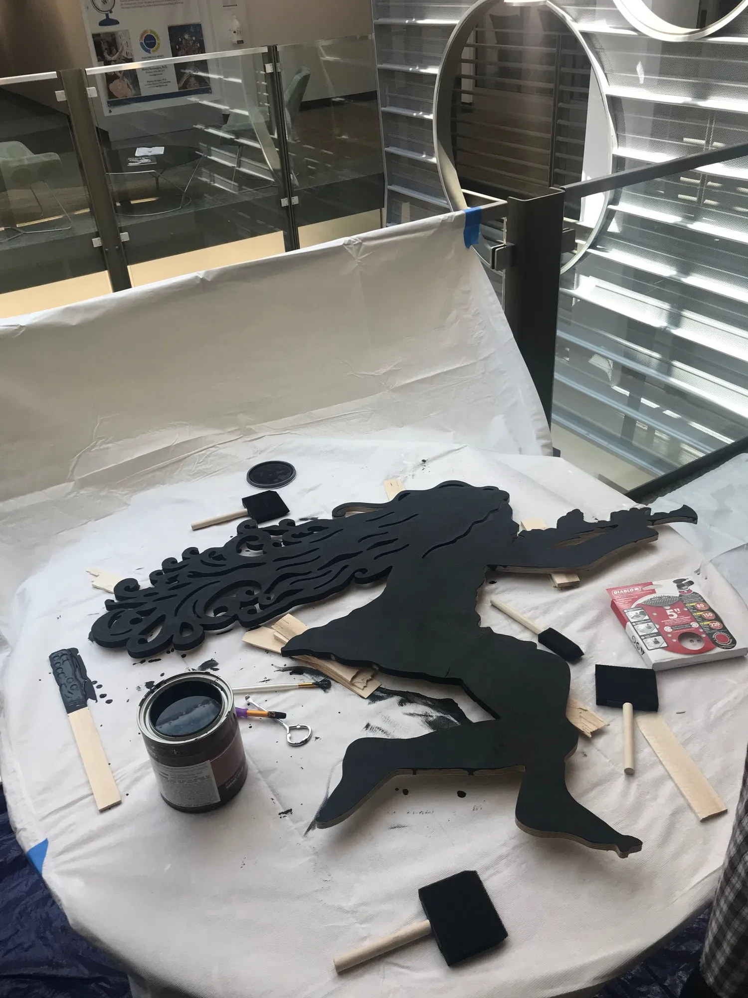 The flute-girl figure fully painted black, laid flat on a table with an open paint can beside her, the elaborate CNC-carved flowing hair and full body now in matte black, ready for installation
