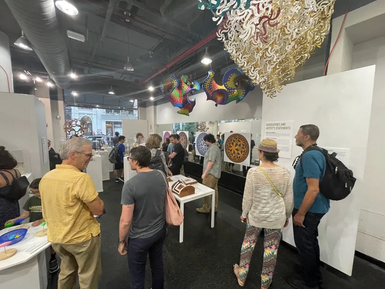 Opening reception: visitors browse prints and sculptures beneath colourful Nico Belmonte hyperbolic ceiling pieces, with the Curvahedra visible through the window