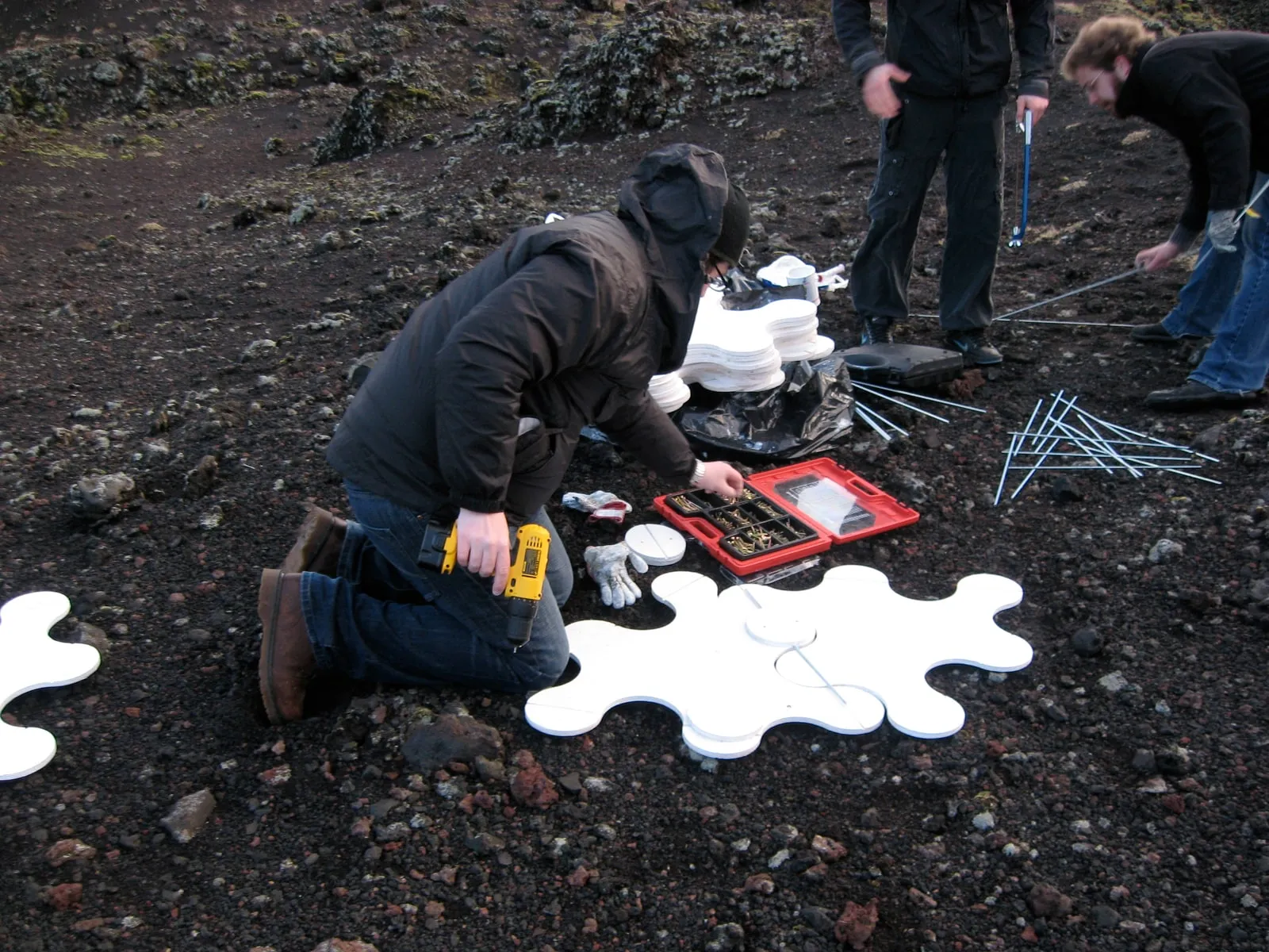 Assembling pieces on the lava field, kneeling with drill and toolbox, the black lava behind