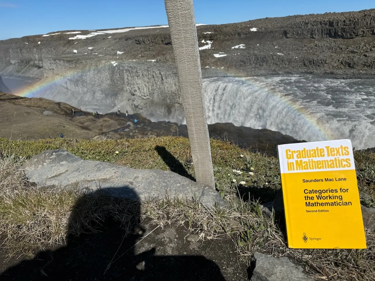 The book on a rock at Dettifoss, a rainbow arcing across the gorge and the photographer's shadow on the foreground stone