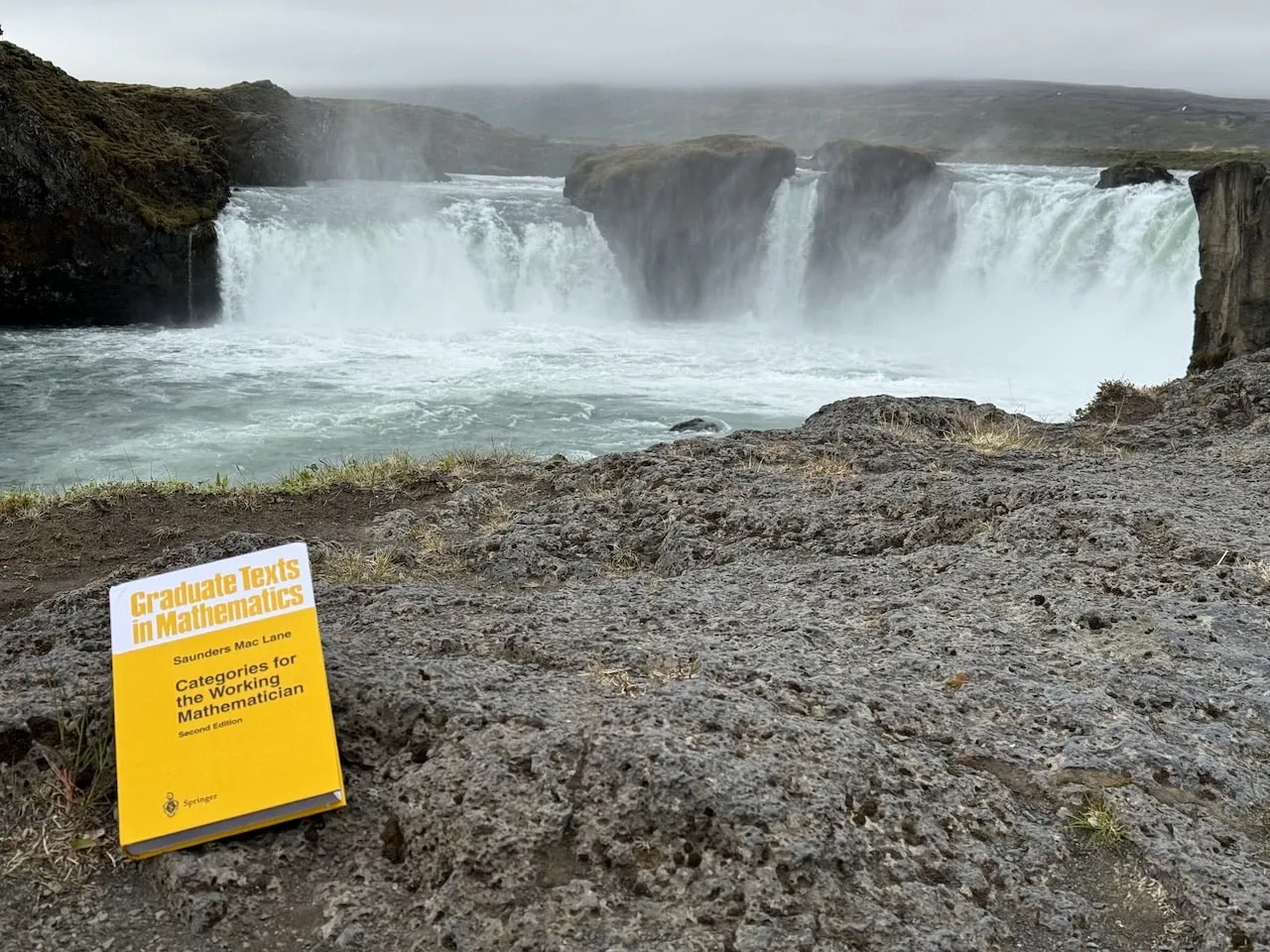 The book on dark rock in the foreground with the wide curved cascade of Goðafoss waterfall thundering behind under low mist