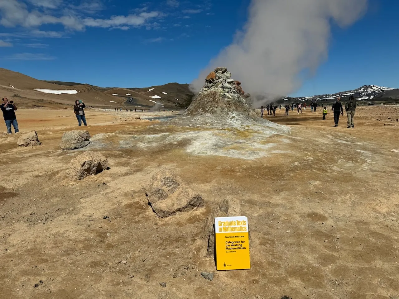 The book on baked orange ground with a steaming cone-shaped fumarole venting in the middle distance of the Hverir geothermal area