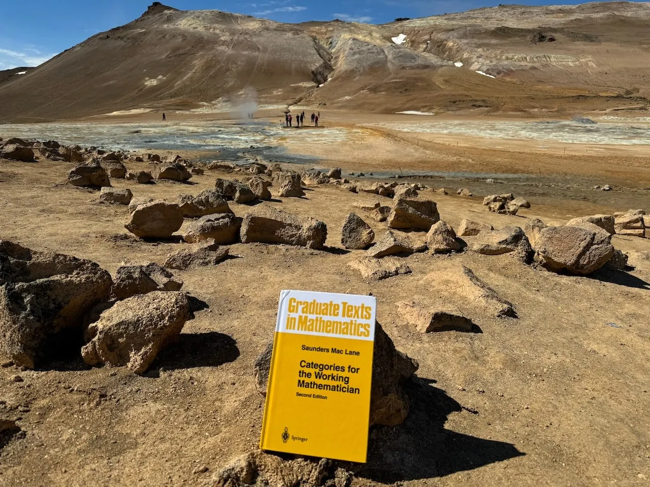 The book standing in cracked ochre earth at the Hverir geothermal field near Mývatn, a steaming mineral-streaked slope and small figures behind
