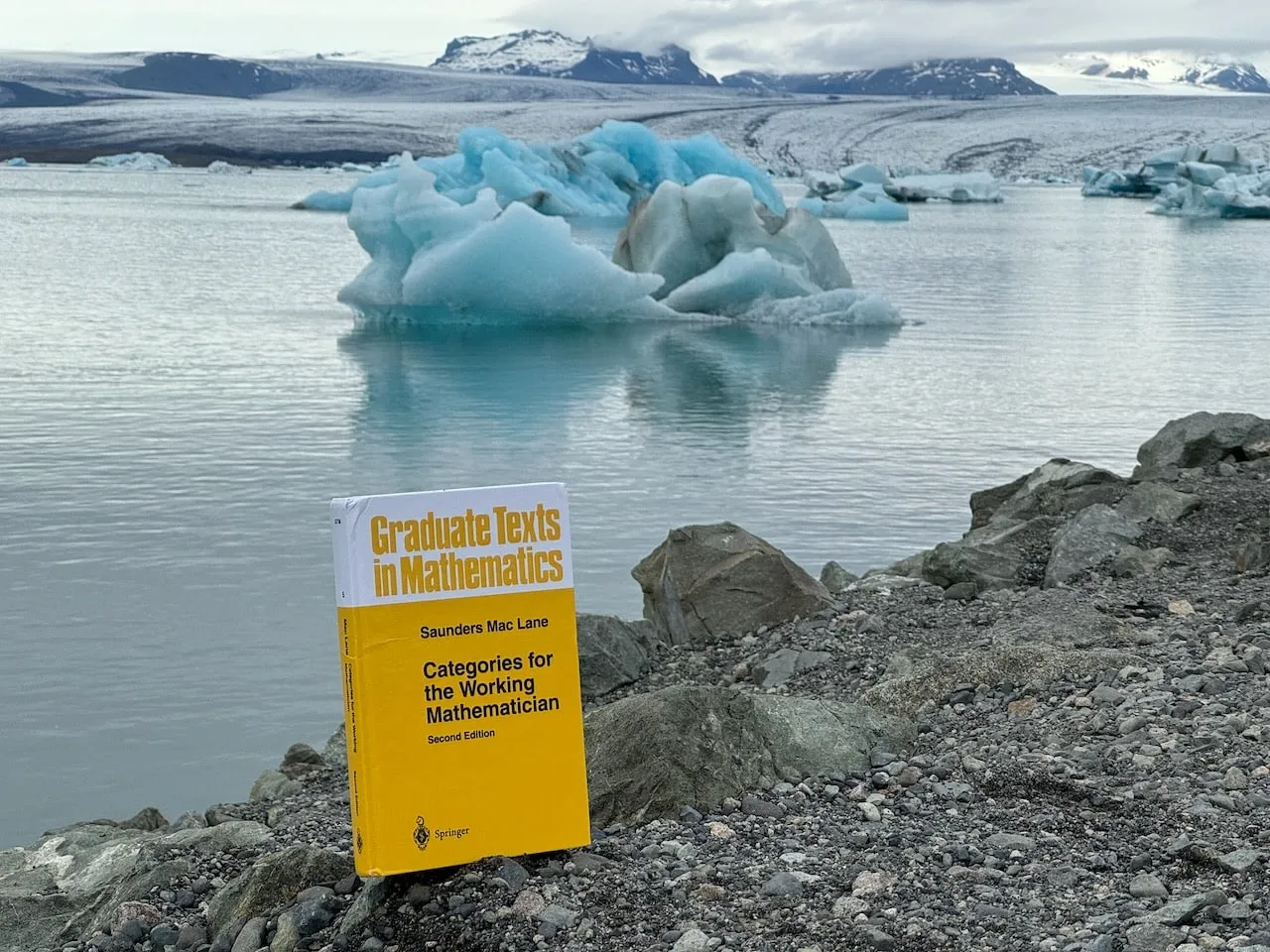 The book on grey rock at Jökulsárlón, a vivid blue iceberg floating offshore on the calm lagoon with snow-streaked mountains behind