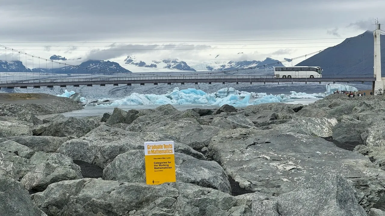 The book among black boulders at Jökulsárlón, blue icebergs drifting beneath the road bridge with a coach crossing and mountains behind