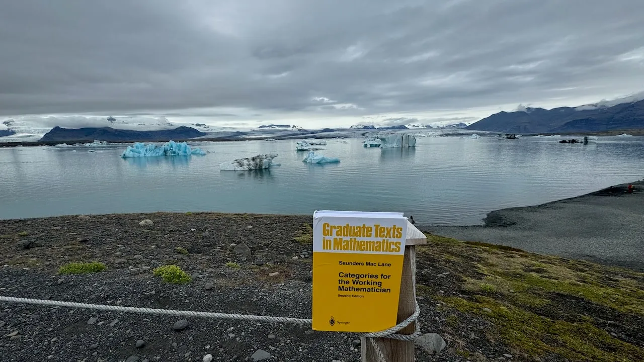 The book balanced on dark rock at Jökulsárlón glacier lagoon, a pale blue iceberg on still water and snow-capped mountains beyond