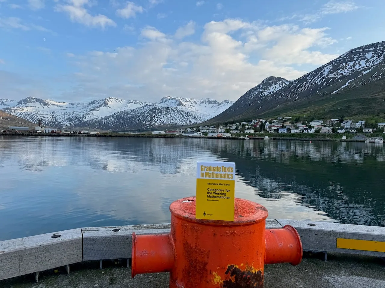 The book balanced on a bright orange mooring bollard at Siglufjörður harbour, the reflective fjord water, waterfront houses and snow-capped peaks behind