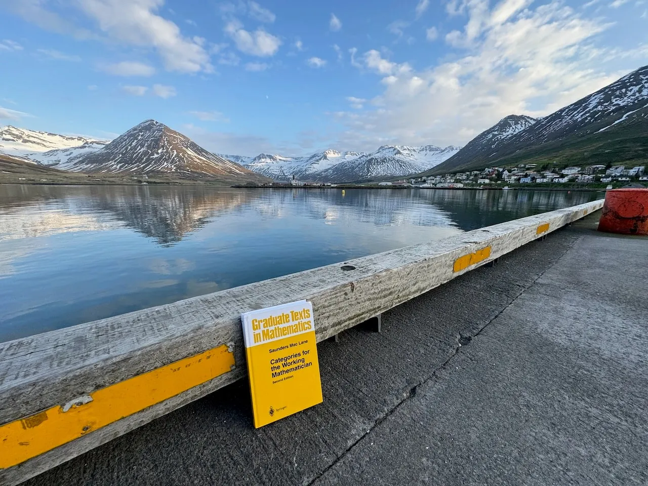 The book propped on a weathered wooden harbour railing at Siglufjörður, the still mirror-calm fjord backed by snow-streaked mountains and the town