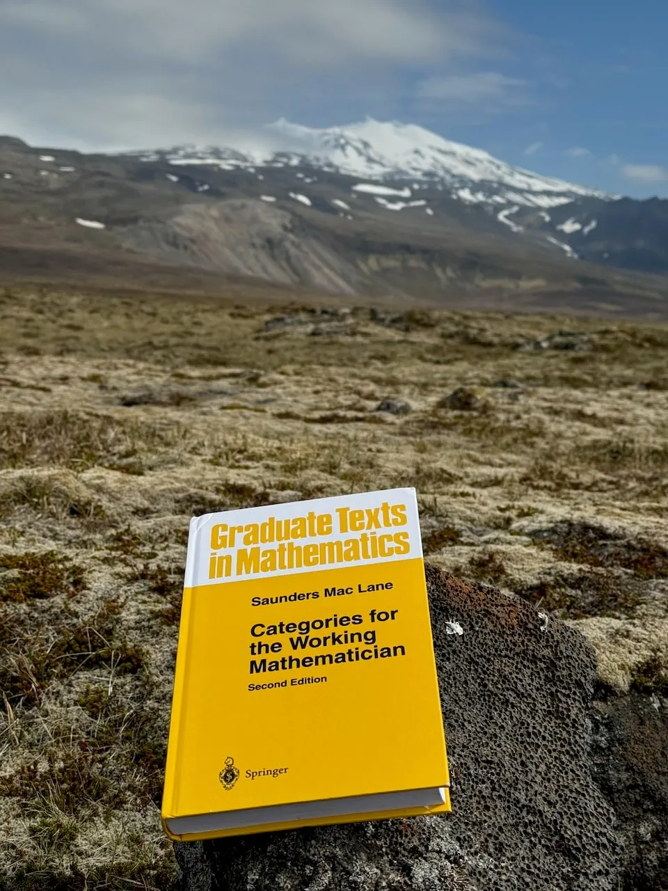 The book standing on lichen-covered rock in a mossy lava field, the snow-capped cone of Snæfellsjökull rising under a clouded sky